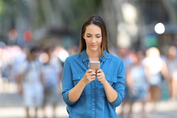 istock Front view of a woman using a smart phone walking on the street 1003787780