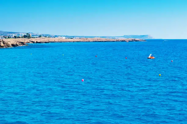 Image of calm sea surface near Agia Napa, Cyprus. One orange paddleboat in deep blue water against rocky coast and cape Greco on the background. Warm day in fall. Concept of leisure and recreation