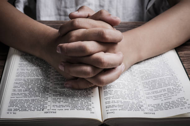 istock Praying hands with opened holy bible on the wooden desk 1003824092
