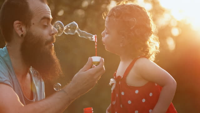 SLO MO Toddler girl blows bubbles into father's face in the setting sun