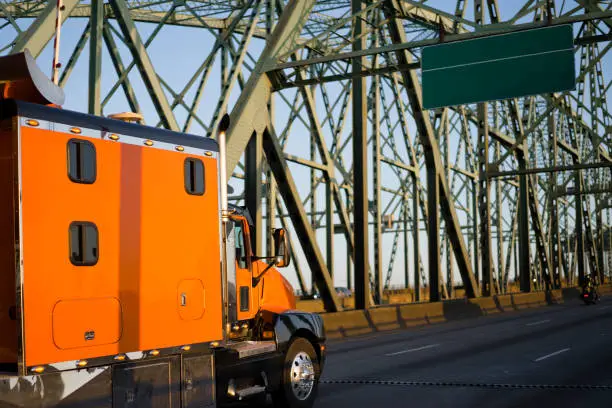 Orange big rig long haul semi truck with extra long cabin for truck driver rest driving alone interstate highway transporting commercial cargo moving on arched metal truss Columbia Interstate Bridge