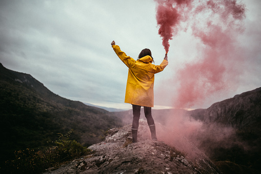 One woman, standing on mountain peak on a rainy day in yellow raincoat, rear view, holding a flaming torch.