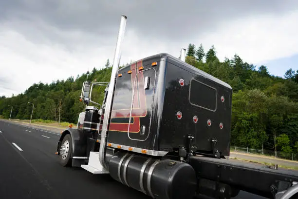 Black classic American bonnet powerful stylish big rig semi truck with vertical exhaust pipes and chrome accent parts on cab with sleeping place for truck driver rest going on wide highway with trees