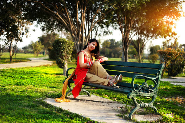 Young woman of Indian ethnicity sitting on public park bench during sunset time, she also looking at camera.