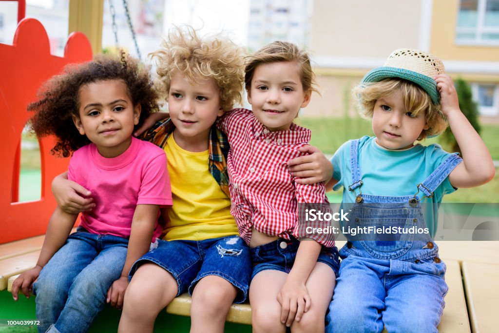 adorable curly boy embracing two multicultural children while other boy sitting near at playground Preschool Age Stock Photo