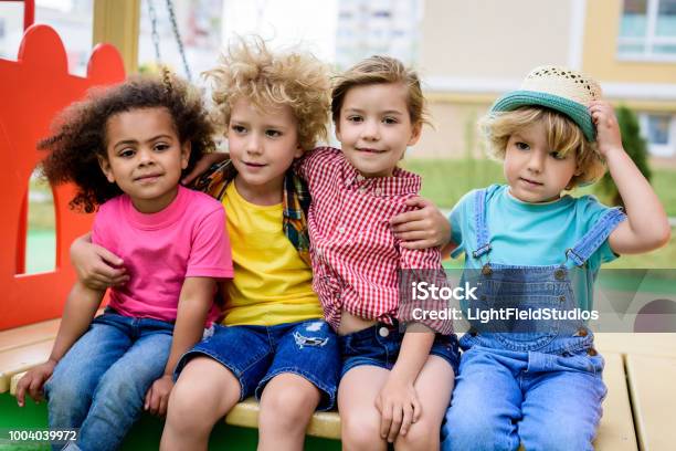 Adorable Curly Boy Embracing Two Multicultural Children While Other Boy Sitting Near At Playground Stock Photo - Download Image Now