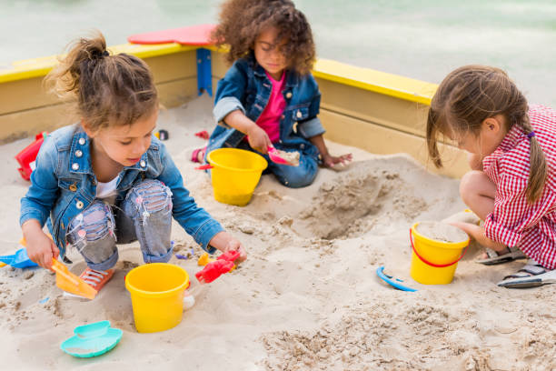 três criancinhas multiétnicas, jogando com colheres plásticas e baldes na areia no playground - preschool child preschooler multi ethnic group - fotografias e filmes do acervo