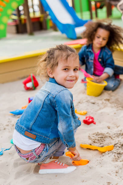 istock selective focus of smiling little child sitting in sandbox with plastic scoop while her friend sitting behind at playground 1004040810