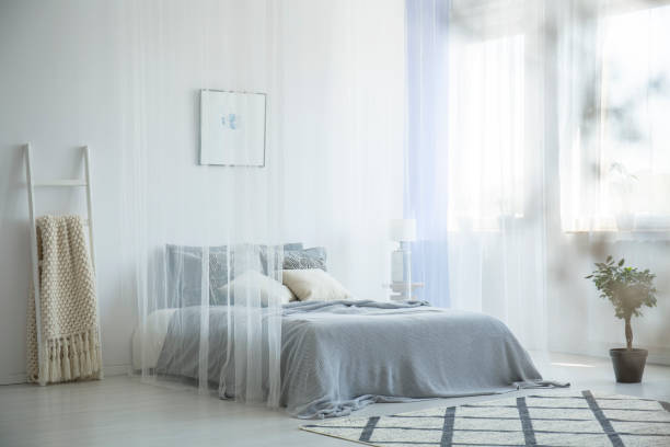 istock Grey knit blanket placed on the bed with canopy in white hotel room interior window with drapes, patterned carpet and decorative ladder 1004109236