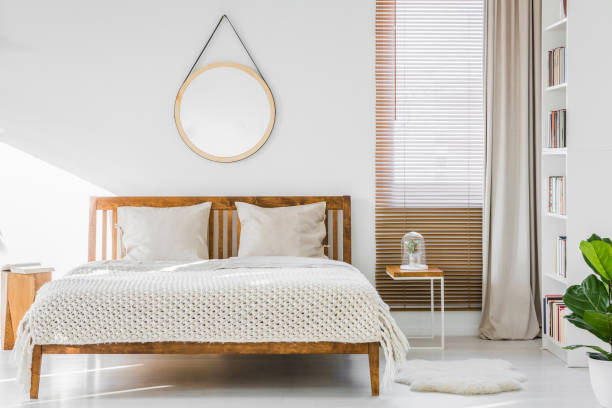 istock Fluffy rug placed next to double bed with white sheets and knit blanket standing in bright room interior with books on white rack, window with wooden blinds and empty round poster on the wall 1004109292