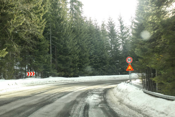 driver view on sharp road curve, partially covered with snow in coniferous forest, with strong blinding sun light. speed limit 50 and bumps sign to illustrate dangerous driving conditions in winter. - skidding bend danger curve imagens e fotografias de stock