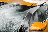 Detail on dark yellow car windshield ad side mirror being washed with water jet spray, cleaning soap foam.
