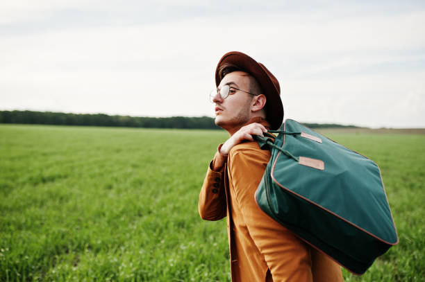 Young man with head down and backpack on the shoulder walking beside wire fence. Rear view of man