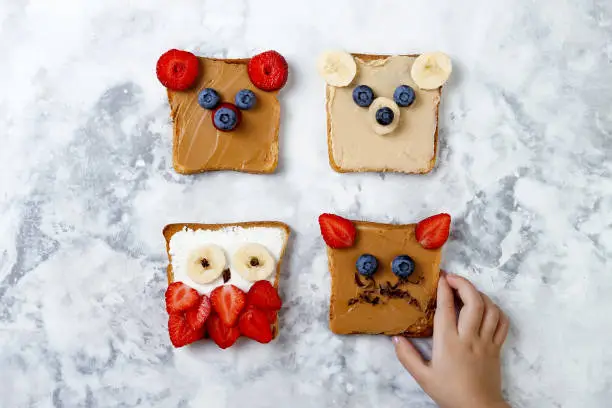 Photo of Healthy funny face sandwiches for kids. Animal faces toast with peanut and cashew butter, ricotta cheese, banana, strawberry and blueberry