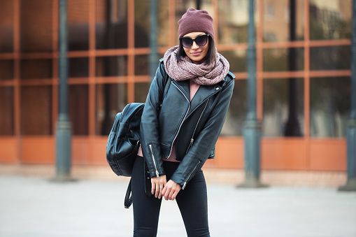 Young fashion woman with backpack walking in city street Stylish female model in leather jacket and black jeans outdoor