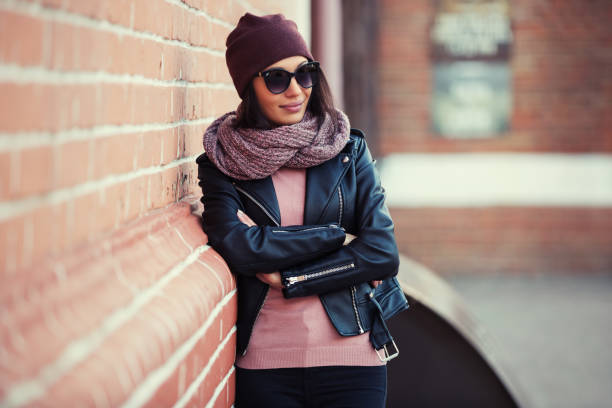 Young fashion woman in black leather jacket and sunglasses leaning on brick wall stock photo