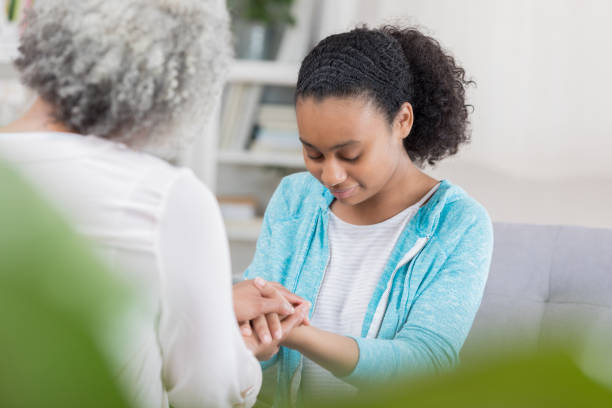 Teenage girl prays with grandmother at home A teenage girl sits on a couch in her living room across from her unrecognizable grandmother. Her head is bowed, eyes closed and she is holding both of her hands. teenage girl with grandmother stock pictures, royalty-free photos & images
