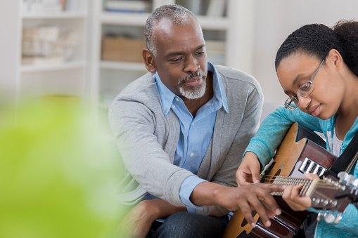 A serious senior man sits at home with his granddaughter.  She holds a guitar and looks down as he reaches forward to point to a fret.