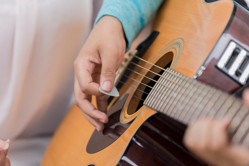 In this closeup, human hands can be seen playing a guitar.  Focus is on the strumming hand holding a pick.  Another hand can be seen  on the fret board chording.