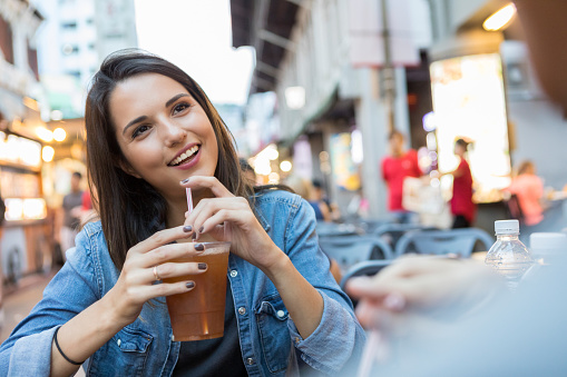 A smiling young woman holds a drink as she sits at a sidewalk cafe in Singapore.  She is looking at her unrecognizable date across the table.