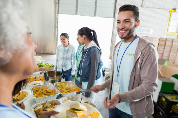 Young man serves senior woman during food bank charity drive A smiling young man stands in a serving line at a food bank and serves a sandwich to an unrecognizable senior woman. soup kitchen stock pictures, royalty-free photos & images