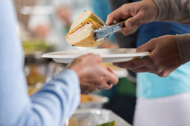 Unrecognizable food bank volunteer serves customer The hands of a food bank volunteer can be seen serving a sandwich plate with tongs to the hands of a customer.  A table of food can be seen in the background. soup kitchen stock pictures, royalty-free photos & images