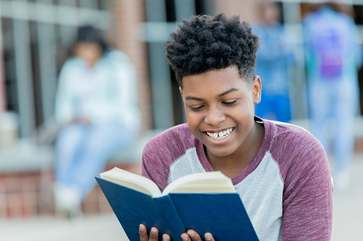 Happy African American male high school student reads book while waiting for school to start.