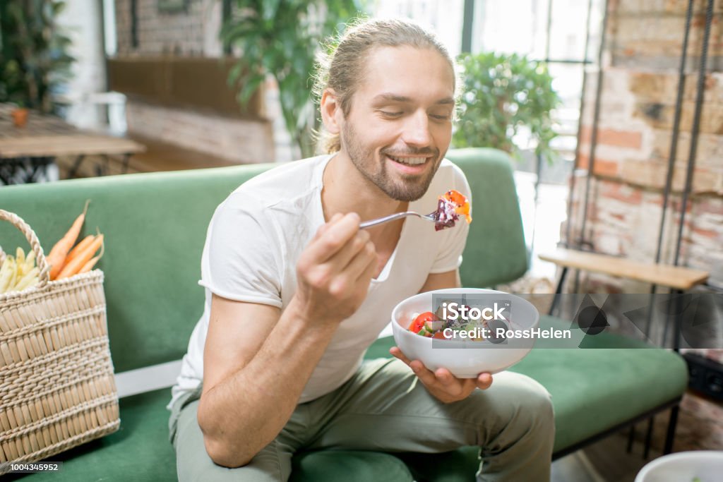 Vegetarian man eating salad indoors Handsome man eating healthy salad sitting indoors on the green sofa with bag full of vegetables on the background. Healthy eating concept Eating Stock Photo