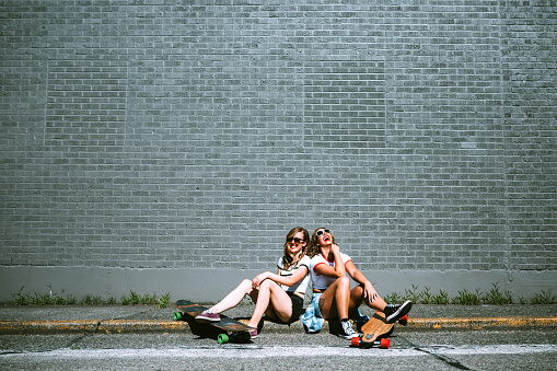 Two young adult women enjoy the summer sun in their town with longboard skateboards.  They sit on the curb in front of a large gray brick wall.