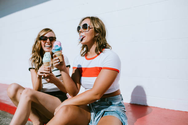 Summer Friends Enjoy Ice Cream Cones on Hot Day Two young adult women have fun eating ice cream cones outside on a warm day of summer vacation. licking ice cream cone stock pictures, royalty-free photos & images