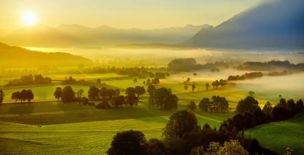 Breathtaking morning landscape of small Bavarian village covered in fog. Scenic view of Bavarian Alps at sunrise with majestic mountains in the background. Breathtaking morning landscape of small Bavarian village covered in fog. Scenic view of Bavarian Alps at sunrise with majestic mountains in the background, Anger, Germany. luz solar stock pictures, royalty-free photos & images