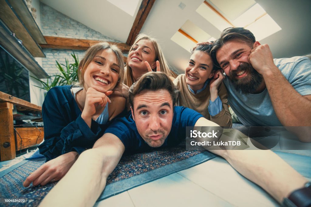 Make a face Five friends, two guys and three girls are lying down on floor one on other. They are taking selfie. One guy is holding camera and they are posing for selfie photo. This people are best friends  and they are spending a lot of time together. Everybody are dressed casual and comfortable. Apartment Stock Photo