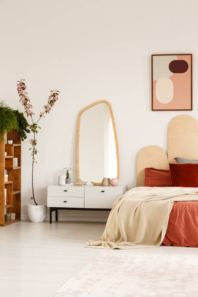 istock Mirror on white cabinet next to bed with red pillows in bright bedroom interior with poster. Real photo 1004567100