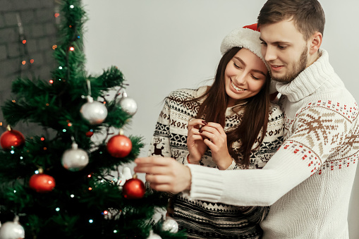 Beautiful woman in santa hat and handsome man in white sweater decorating green christmas tree with ornaments and lights, romantic couple greeting card concept
