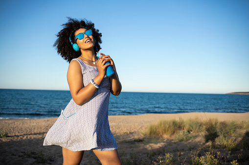 Young african woman with colorful headphones having fun and drinking cocktail on beach.