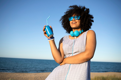 Young african woman with colorful headphones having fun and drinking cocktail on beach.