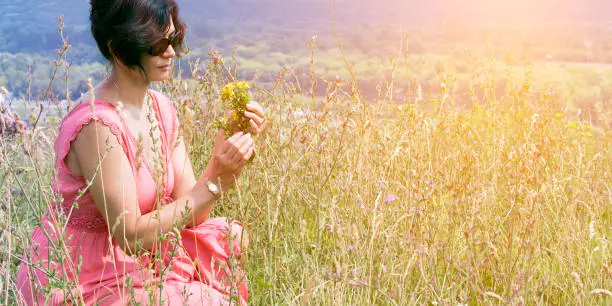 A middle-aged woman in a pink dress collects medicinal herbs in a meadow.