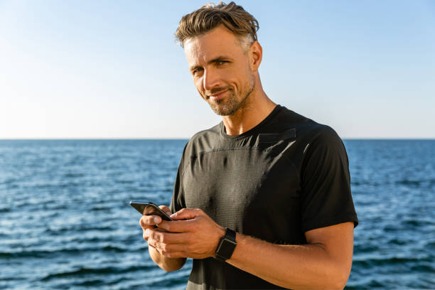 istock handsome adult man with smartphone on seashore and looking at camera 1004830946