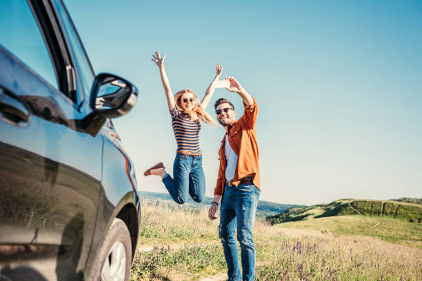 happy young woman jumping with wide arms while her boyfriend taking selfie on smartphone near car in field happy young woman jumping with wide arms while her boyfriend taking selfie on smartphone near car in field hopper car stock pictures, royalty-free photos & images