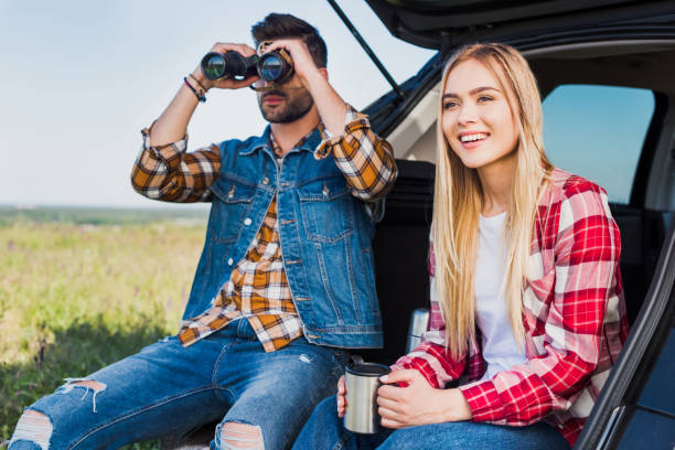 istock male tourist looking through binoculars while his smiling girlfriend sitting near with coffee cup on car trunk in field 1004889840