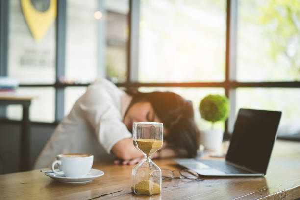 freelancer asian businesswoman tired after working coffee shop her sleeping on workplace table near windows and hourglass at evening with digital laptop computer and coffee break. - waiting women clock boredom imagens e fotografias de stock