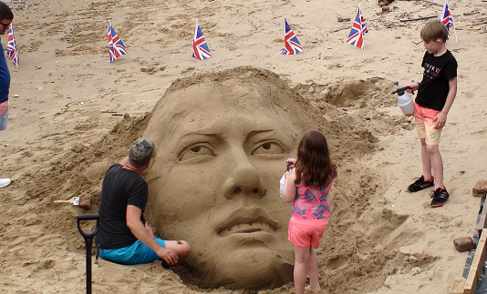 View Of British Flag Waving And A Man Sitting On The Ground, Creating A Face Sculpture With Sand While Kids Are Standing, Holding Water Plastic Jar And Watching During The Day At South Bank Of Thames River In Westminster London England Europe