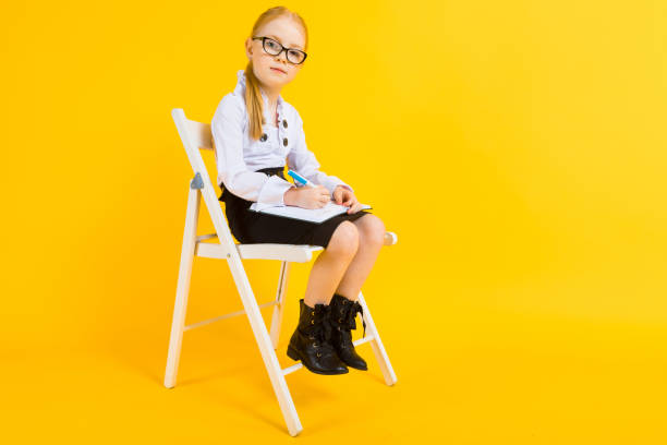 Girl with red hair on a yellow background. A charming girl in transparent glasses sits on a white chair and makes notes in a notebook. Portrait of a beautiful girl in a white blouse and black skirt. see through leggings stock pictures, royalty-free photos & images
