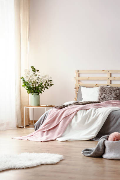 istock Fresh plants in ceramic vase standing on bedside table in white bedroom interior with window with curtains, king-size bed and white fluffy rug on the floor 1005124648