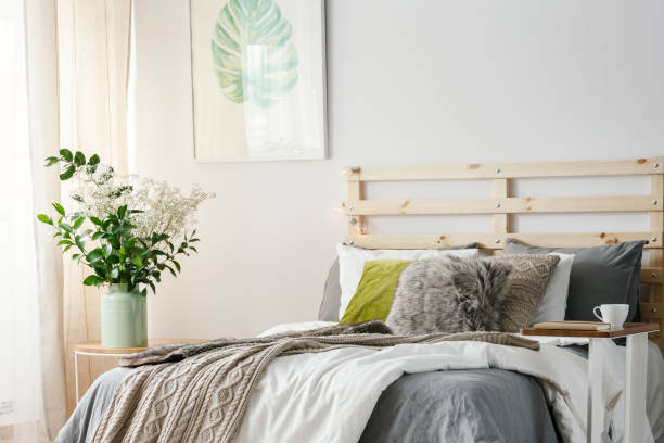 istock Small table with notebook and coffee cup standing by the king-size bed with blanket and fur pillow in bright room interior with poster and flowers in vase 1005124652