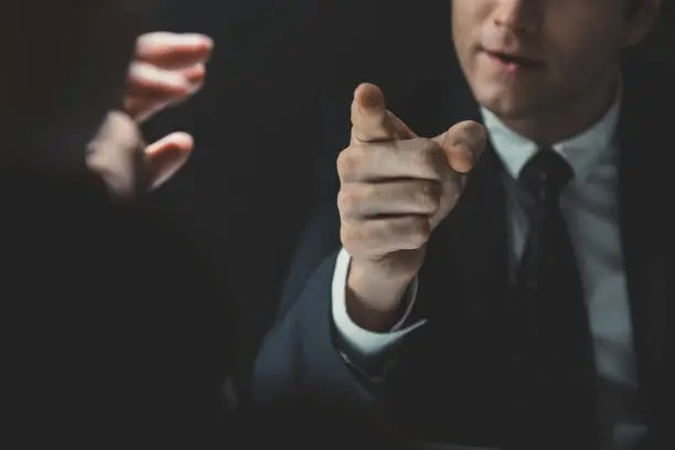 Angry man in formal business suit pointing hand to somesone he talking to in dark interrogation room