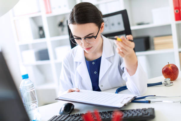 une belle jeune fille dans une robe blanche est assis à un bureau d’ordinateur avec des documents et un stylo dans ses mains. - patient hospital doctor nurse photos et images de collection