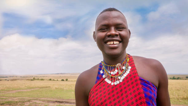 eine nahaufnahme eines lächelnden jungen masai mannes trägt die traditionelle rote kleidung und schmuck seines stammes. die landschaft der masai mara national reserve im hintergrund. - costume expressing positivity cheerful close up stock-fotos und bilder