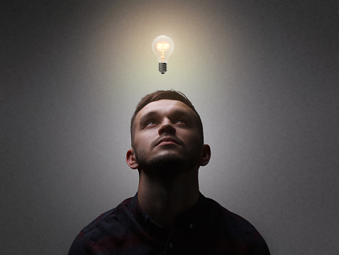 New idea, innovation, and creativity concept. Close up portrait of a man which looks up getting an idea represented by a light bulb overhead. Grey background, copy space.