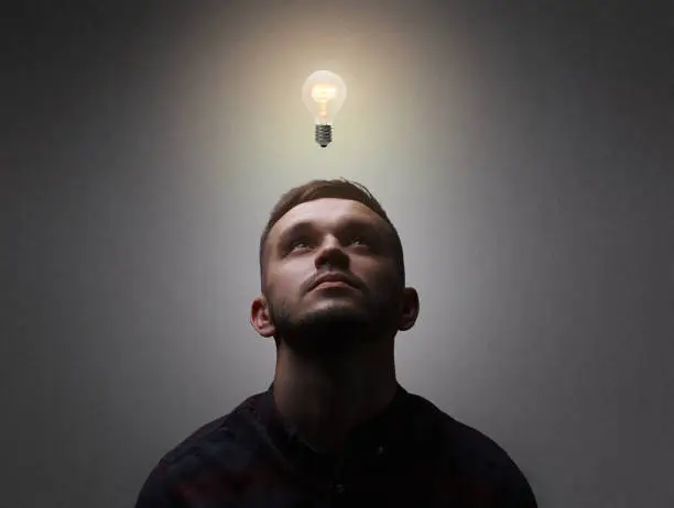 Photo of New idea, innovation, and creativity concept. Close up portrait of a man which looks up getting an idea represented by a light bulb overhead. Grey background, copy space.